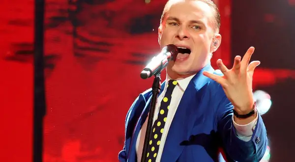 Ralph Kaminski w niebieskiej marynarce i krawacie w grochy śpiewa do mikrofonu. Jego płyty to muzyczna podróż.