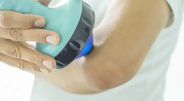 Une main utilise un appareil bleu clair pour soigner une tendinite au coude. La lumière bleue émise par l'appareil cible la zone douloureuse.