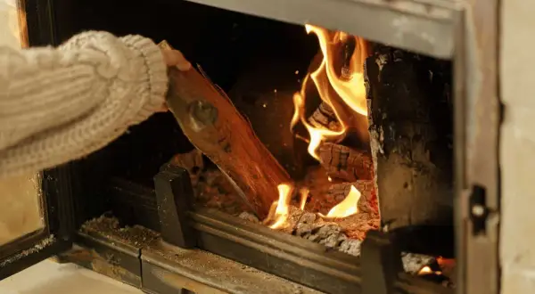 Main tenant ajoutant une bûche dans un poêle à bois, les flammes dansent joyeusement.
