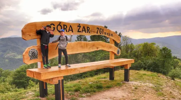 Dzieci cieszą się na ławce z napisem "Góra Żar 761 m n.p.m.". To świetne atrakcje dla dzieci w górach.