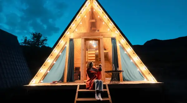Soirée de camping nature corse. Une femme assise sur les marches d'un chalet en bois illuminé par des guirlandes lumineuses, profitant de la nuit.