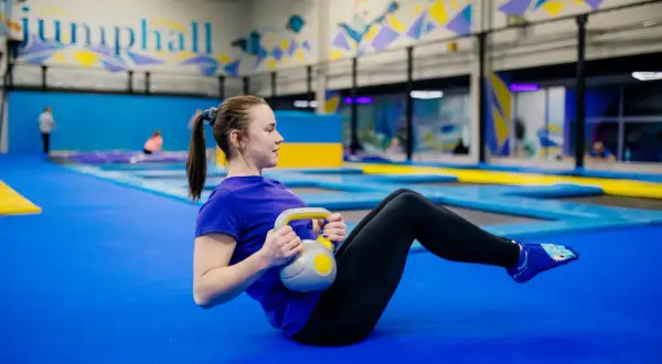 Kobieta ćwiczy z kettlebell na niebieskiej macie w sali gimnastycznej z trampolinami w tle.