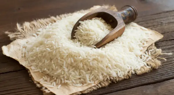 Ein Holzlöffel schöpft weißen Langkornreis aus einem Haufen auf Jute. Der Unterschied zwischen Basmati-Reis und Langkornreis ist hier visuell dargestellt.