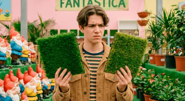 Joven indeciso compara dos muestras de césped artificial, buscando el mejor césped artificial del mercado. Detrás, gnomos de jardín y plantas.