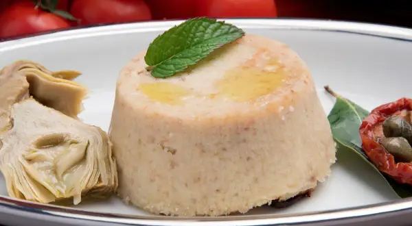 Delizioso flan di carciofi, guarnito con menta e accompagnato da carciofi sott'olio, pomodori secchi e capperi.