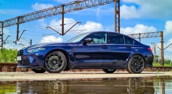 Granatowe BMW M3 G80 stoi obok torów kolejowych, odbijając się w kałuży.