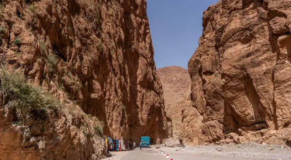 Die Todra-Schlucht mit ihren steilen, roten Felswänden. Eine Straße führt hindurch, gesäumt von Souvenirständen und einem blauen LKW.