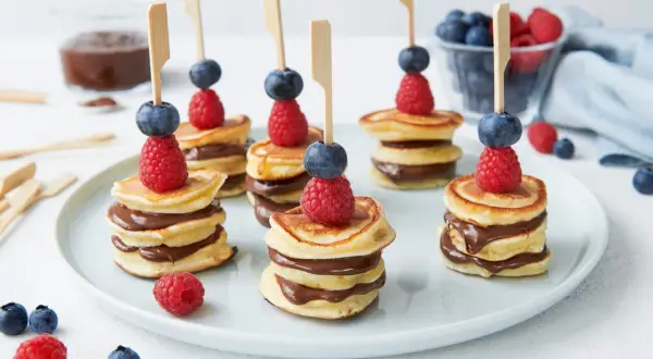 Mini pancakes rezept: gestapelte Mini-Pfannkuchen mit Schokocreme, garniert mit Himbeeren und Blaubeeren auf Spießen.