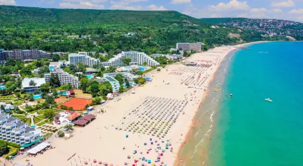 Słoneczne bułgarskie plaże tętnią życiem. Widać hotele, mnóstwo leżaków i parasoli, a także turystów kąpiących się w turkusowym morzu.