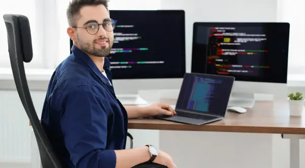 Typowy programista w okularach siedzi przy biurku z trzema monitorami wyświetlającymi kod.