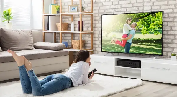 Une jeune femme se repose sur un tapis blanc, télécommande en main, pour regarder la tv sans connexion internet. L'écran affiche un couple s'enlaçant.