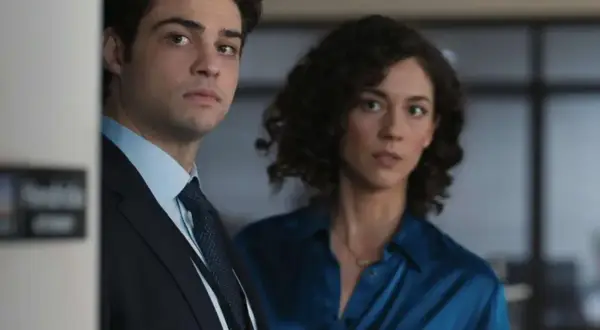 Besetzung von The Recruit: Ein Mann im Anzug und eine Frau in einem blauen Hemd blicken ernst in die Kamera.