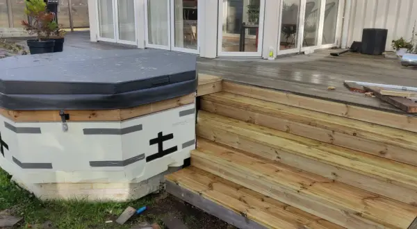 Pågår bygga badtunna. Isolerad stomme med trädetaljer och en grå presenning. Nya trätrappor leder upp till altanen.