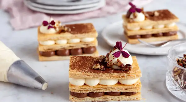 Tre eleganta lager av smördeg, chokladkräm och vaniljkräm toppas med nötter och en lila blomma. En lyxig dessert redo att avnjutas.