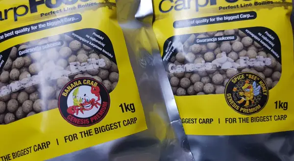 Dwie paczki z żółtymi etykietami zawierają kulki na karpia. Na opakowaniach widnieje napis "CarpFood" i grafika z pszczołą.