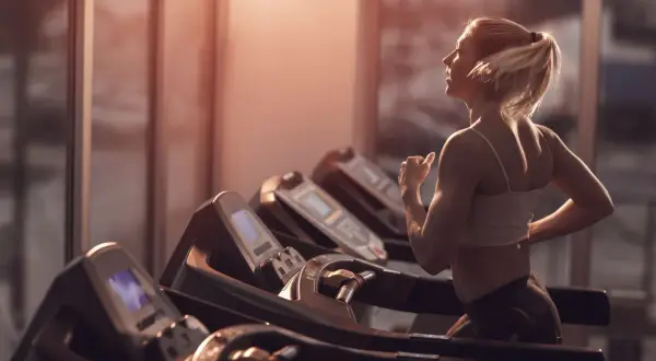 Frau mit blondem Pferdeschwanz walkt auf dem Laufband im Fitnessstudio, das warme Abendlicht fällt durch die Fenster.