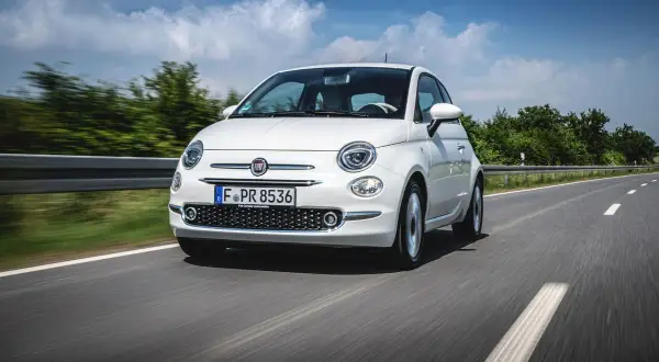 Biały fiat mini pędzi po asfaltowej drodze, otoczony zielenią i błękitnym niebem z chmurami.