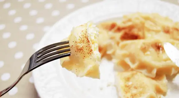 Widok z bliska na widelec z pierogiem z serem, posypanym cynamonem. Na talerzu widać więcej pysznych pierogów.