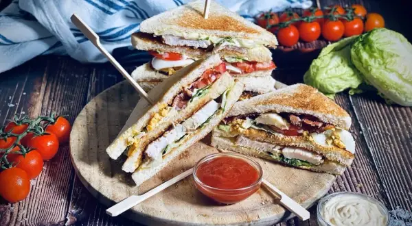 Leckere Sandwich-Ideen: Gestapelte Club-Sandwiches mit Hähnchen, Speck, Ei und Tomaten auf einem Holzbrett, daneben Tomaten und Salat.