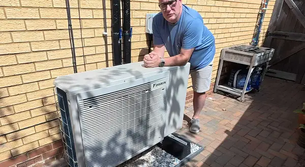 Un homme pose près d'une unité extérieure de pompe à chaleur Daikin, un système efficace pour le chauffage et la climatisation, potentiellement alimenté par une énergie solaire.