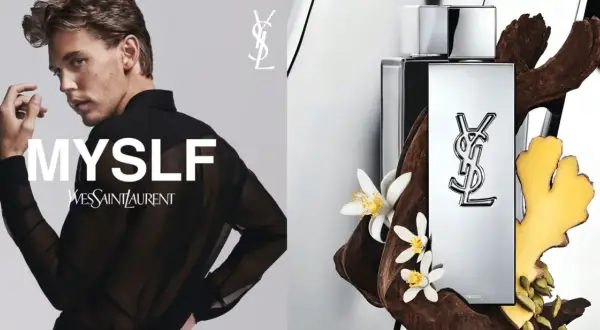 Młody mężczyzna w czarnej, prześwitującej koszuli, obok flakonu perfum Yves Saint Laurent MYSLF, otoczony drewnem i kwiatami.