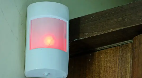Biały czujnik ruchu z czerwoną diodą, zamontowany na ścianie obok drewnianej framugi. Pokazuje, jak działa czujnik ruchu.
