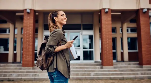 Uśmiechnięta studentka z plecakiem i telefonem przed budynkiem uczelni. Zastanawia się, jakie studia po rozszerzonym angielskim wybrać.