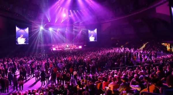 Wielki koncert w Stadionie Wrocław. Tłumy fanów, scena z orkiestrą i ekranami, na których widać dyrygenta.