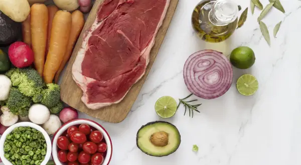 Un corte de carne magra, rodeado de verduras frescas como zanahorias, brócoli, tomates y aguacate, junto a aceite de oliva y limón.