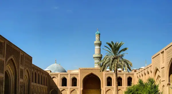 Ein Minarett und eine Kuppel einer Moschee ragen über traditionelle Gebäude mit Bögen und Palmen. Einblick in die reiche irakische Kultur.