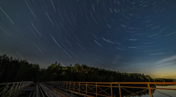 Nocne niebo nad starym mostem, gdzie gwiazdy kreślą na ciemnym tle świetliste ślady.