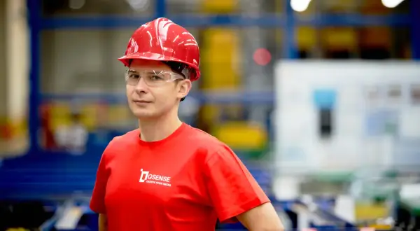 Mężczyzna w czerwonym kasku i okularach ochronnych, w czerwonej koszulce z logo firmy, na tle hali produkcyjnej.
