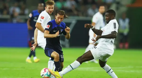Piłkarze walczą o piłkę na boisku, jeden z nich w białej koszulce z logo Bundesligi, być może to przyszli bundesliga mistrzowie.