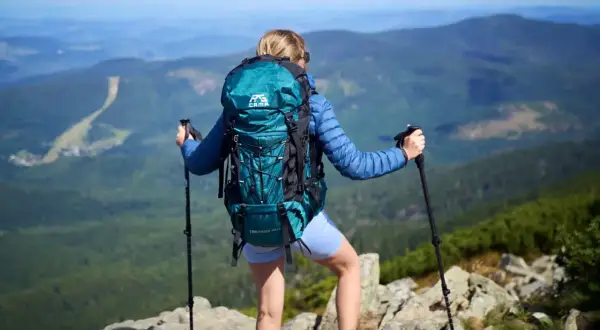Kobieta z turkusowym plecakiem w góry, z kijkami trekkingowymi, podziwia panoramę. Idealny plecak na górskie wędrówki.