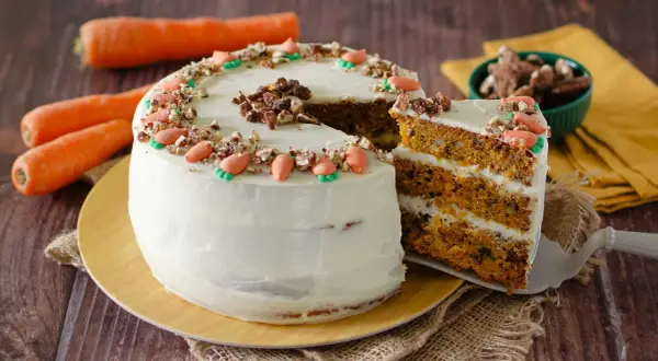 Torta di carote con glassa bianca, decorata con carote di zucchero e noci. Una fetta mostra gli strati soffici, perfetta per una carrot cake ricetta semplice.