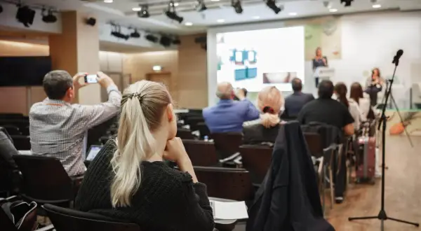 Uczestnicy konferencji, gdzie odbywają się **usługi szkoleniowe**. Jedna osoba filmuje prezentację telefonem.