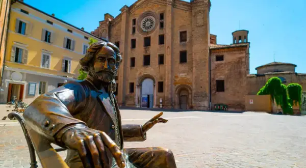 Statua in bronzo di un uomo barbuto, un monumento in Emilia Romagna, con una chiesa in mattoni sullo sfondo.