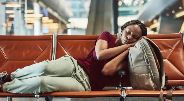 Frau schläft auf einer Bank im Flughafen bei Nacht, ihr Kopf ruht auf ihrem Rucksack.