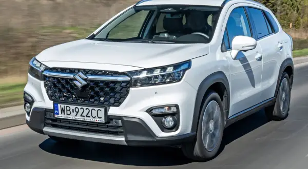 Biały Suzuki S-Cross jedzie po drodze, widoczny przód auta z grillem i światłami.