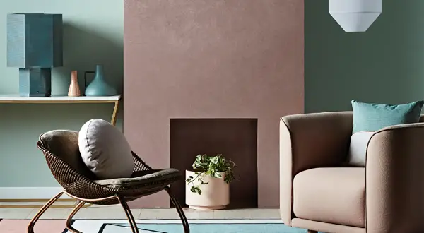 Simulazione abbinamento colori pareti: un salotto con poltrona in vimini, camino rosa e divano beige, su un tappeto geometrico.