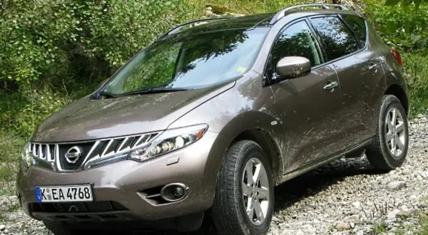 Brązowy Nissan Murano na kamienistej drodze, otoczony zielenią.