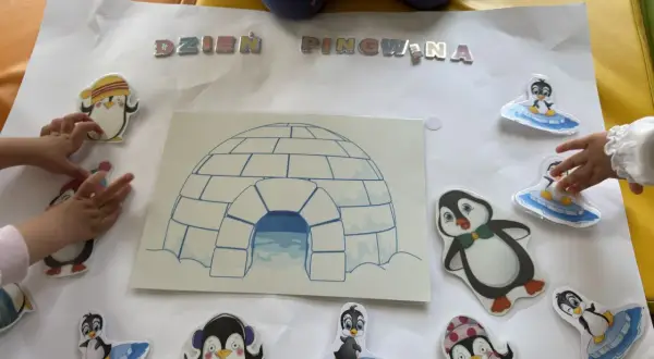 Dzieci tworzą plakat na dzień pingwina w przedszkolu, ozdabiając go naklejkami pingwinów i igloo.