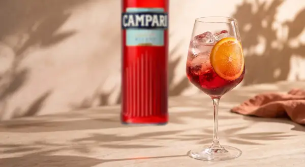 Orzeźwiający Campari Spritz z plasterkiem pomarańczy i kostkami lodu, obok butelki Campari. Idealny na letnie popołudnie.
