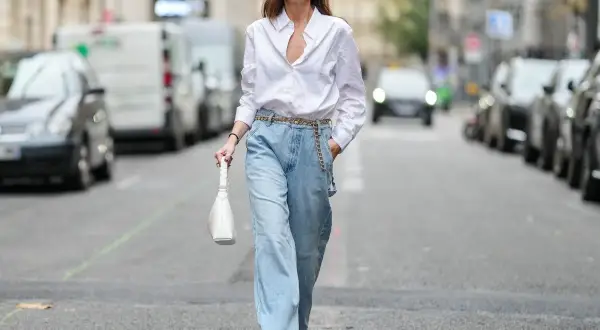 Kobieta w luźnych, jasnych jeansach typu mom fit i białej koszuli, z białą torebką. Idealny look dla każdej kobiety ceniącej wygodę i styl.