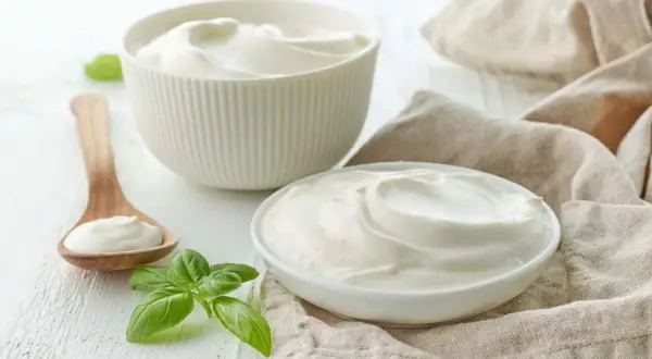 Gładki, kremowy jogurt naturalny w miseczkach i na drewnianej łyżce, ozdobiony listkami bazylii.