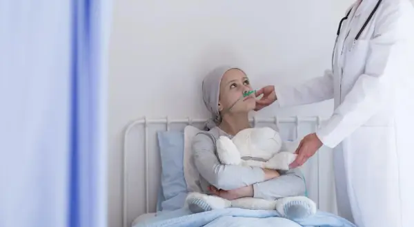 Dziewczynka z rakiem serca w szpitalnym łóżku, z tlenem i pluszowym misiem, otrzymuje wsparcie od lekarki.