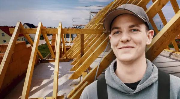 Junger Mann mit Kappe lächelt vor Dachstuhl. Ausbildung Zimmermann Gehalt ist ein guter Start.