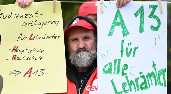 Mann mit Bart und roter Weste hält Schilder hoch. Ein Schild fordert "A13 für alle Lehrer" und thematisiert das Lehrer Gehalt Berlin.