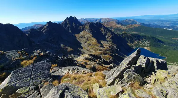 Widok z kamienistego szczytu w Tatrach Wysokich na skaliste granie, trawiaste zbocza i turkusowe jezioro.