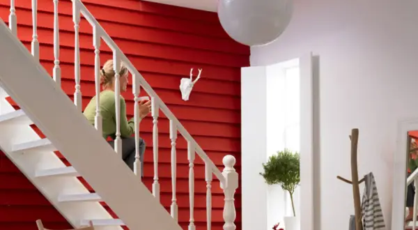 Ein rotes, holzvertäfeltes Treppenhaus mit weißer Balustrade. Eine Frau sitzt auf der Treppe. Ein Hirschkopf und ein Baum schmücken die Wand. So lässt sich ein Treppenhaus gestalten.